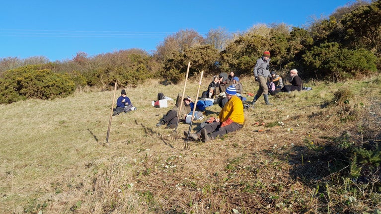 A group of people sit to rest on a sunny slope, after taking part in a scrub bash